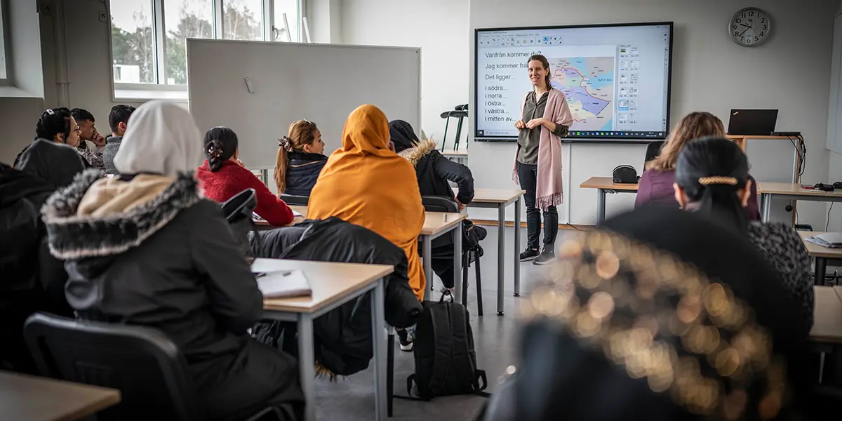 En lärare står vid en tavla längst fram i klassrummet. Det sitter elever och lyssnar på läraren.