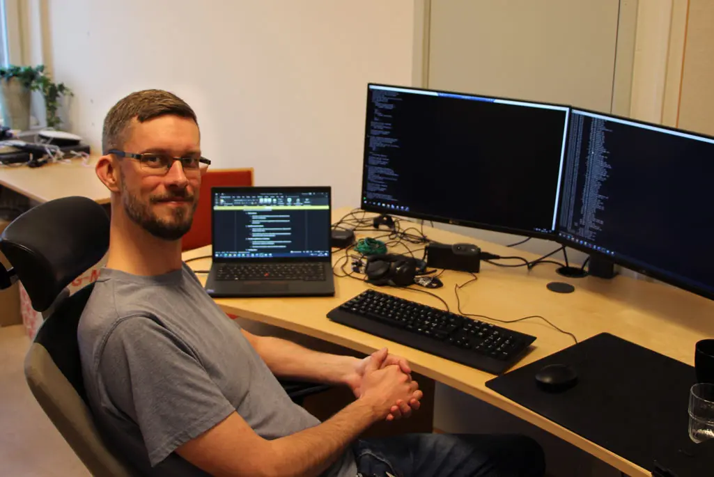 Foto på man med glasögon som sitter på en datastol vid sitt skrivbord. Han är vänd mot kameran och ler.