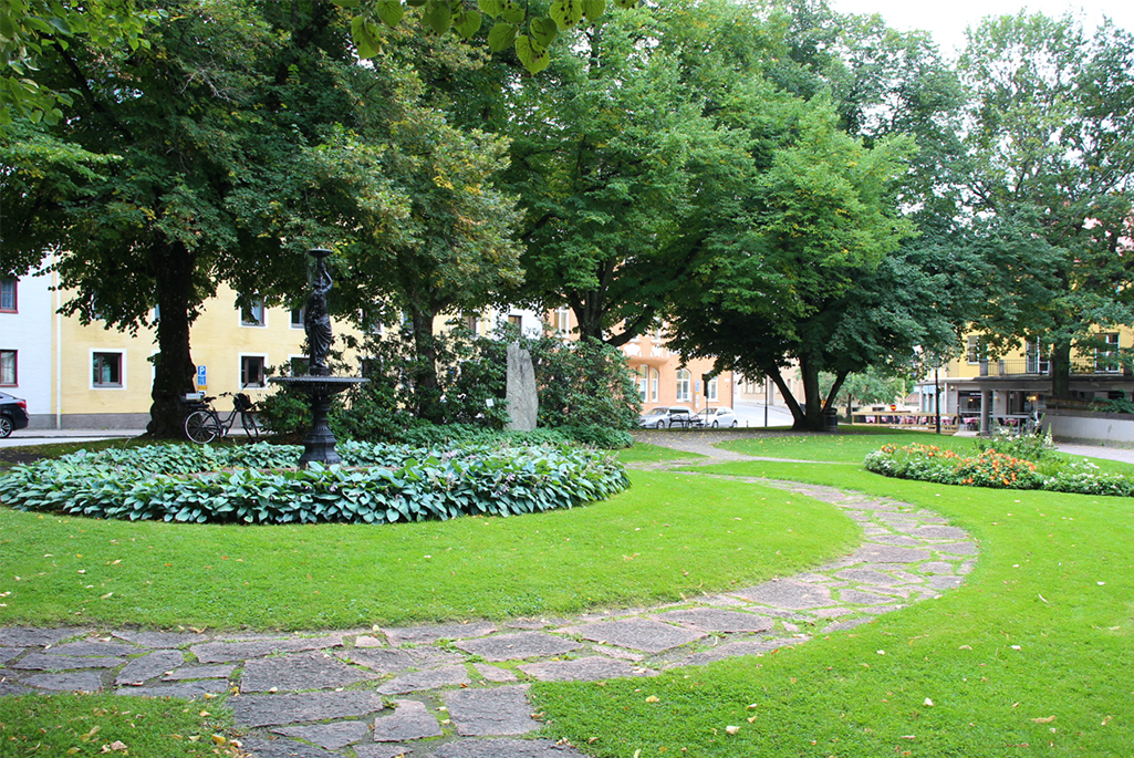 Foto på en park under sommaren. I parken syns en fontän som är omgärdad av perennen hosta. I bakgrunden syns en mindre perennplantering och mellan planteringarna syns en stenlagd stig.