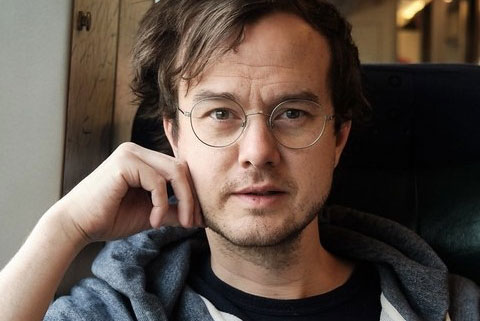 Man med brunt hår och grå tröja, med handen mot kinden, som tittar mot kameran.
