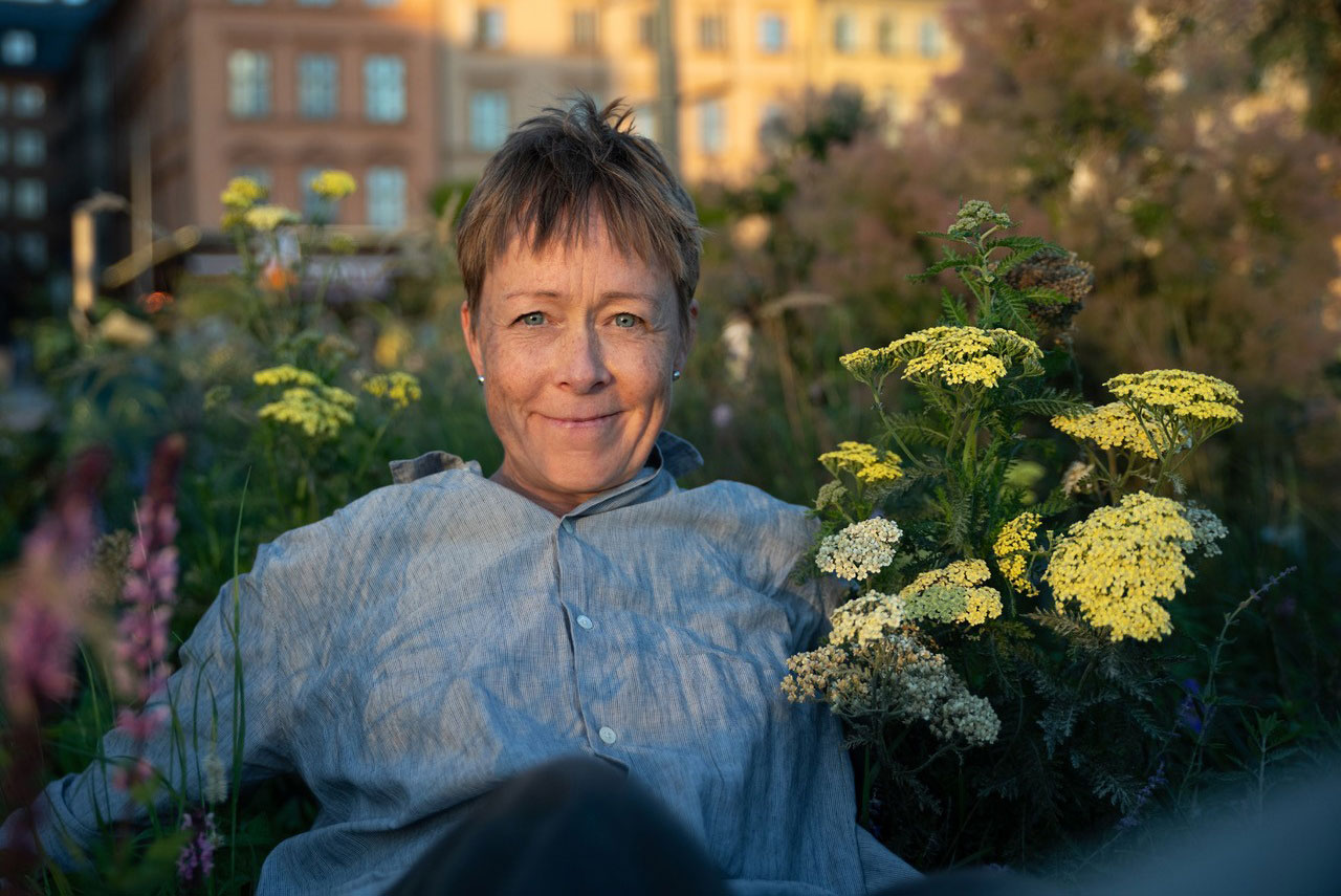 Foto på kvinna som sitter bland grönska och tittar mot kameran.