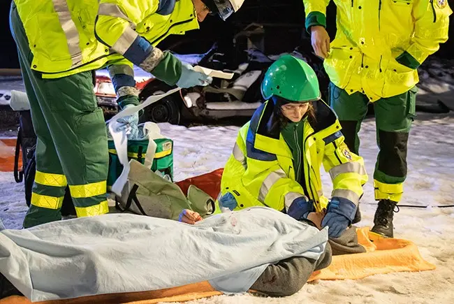 Bild som visar en ung person som utför hjärt- och lungräddning på en docka som ligger på en ambulansbrits utomhus.